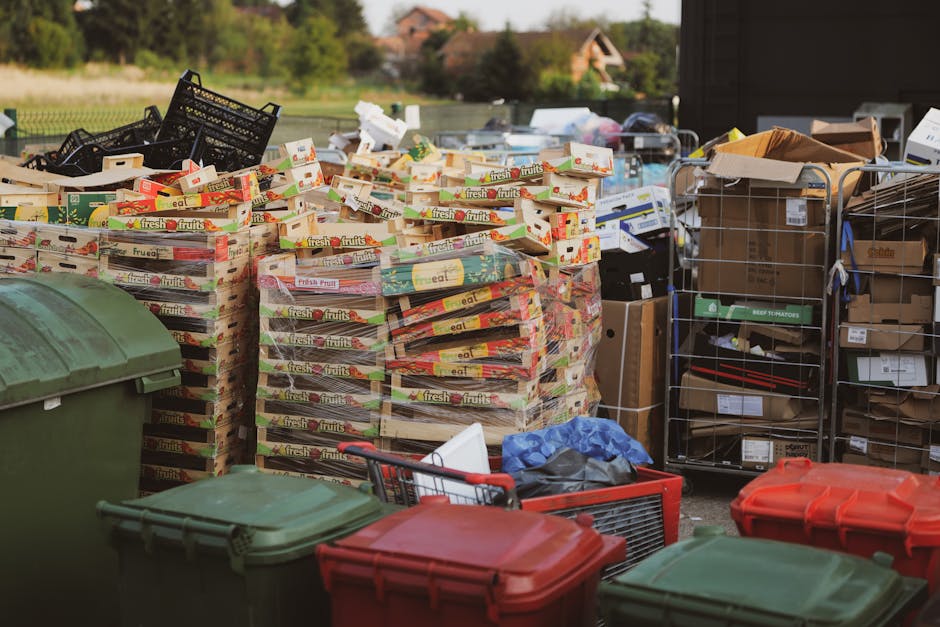 An outdoor scene featuring a large collection of waste and recyclable materials arranged on a paved surface, including multiple stacks of empty fruit and vegetable crate boxes, primarily made of cardb