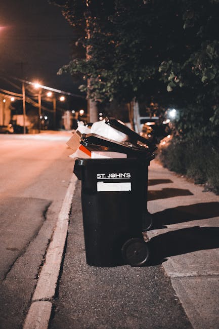 A light grey, rectangular rubbish bin made of smooth plastic, positioned on a wooden surface indoors. The bin is filled with crumpled paper and torn cardboard, with some waste protruding from the open