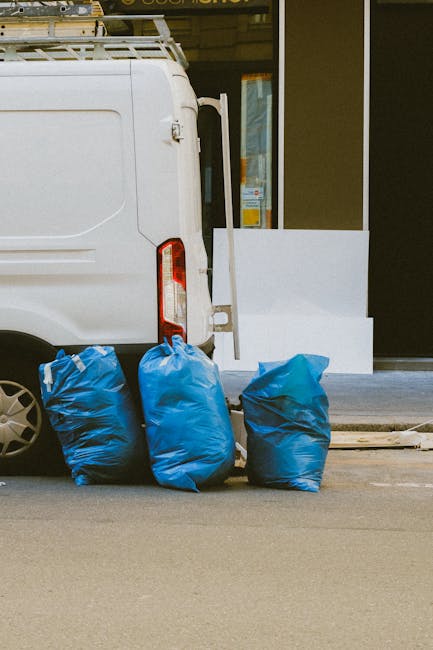 A man seen from the back is walking down a narrow urban street, pushing a hand truck loaded with a large, irregularly shaped black garbage bag secured with black ties. He is wearing a short-sleeved, p