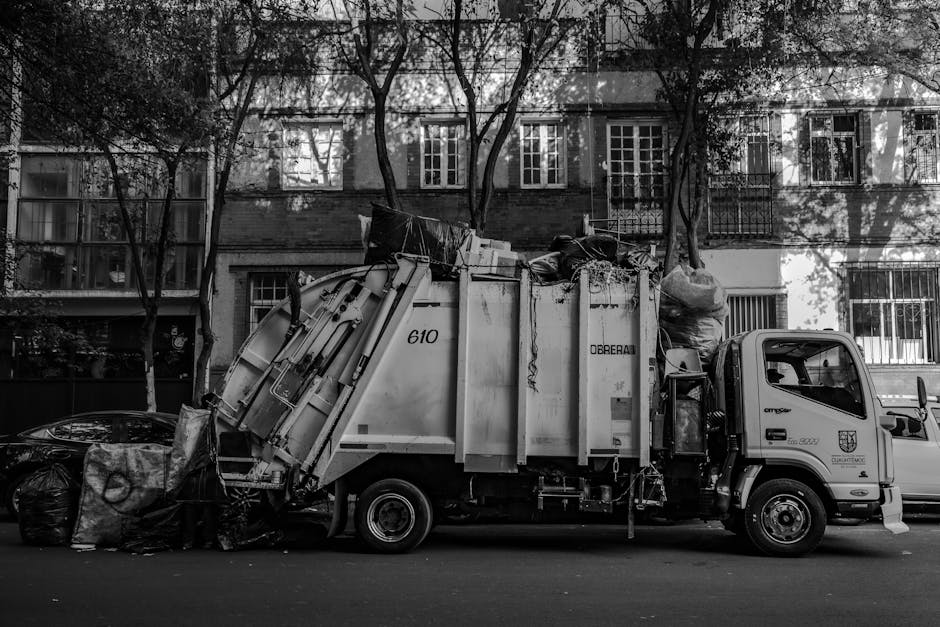 A dark red flatbed utility vehicle parked on an urban street, loaded with various types of waste bags and discarded items, including large black and white garbage bags, some with visible creases and f
